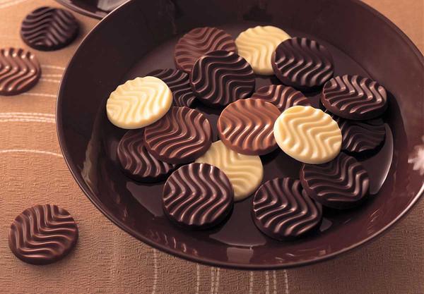 A bowl filled with round chocolate discs featuring a wavy pattern, in dark, milk, and white chocolate varieties, resembling thin cookies.