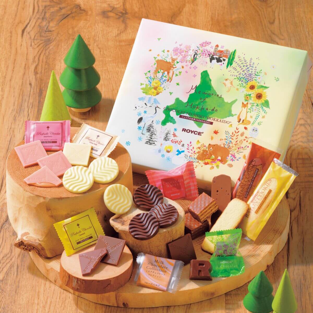 ROYCE' Chocolate - Chocolat No Shiki "Hokkaido" - Image shows wooden stands with confections in different shapes and colors. Accents include miniature green treesand a yellow printed box with text saying Memories of Hokkaido. Chocolat No Shiki "Hokkaido". ROYCE'. Background is in brown with wood texture effect.