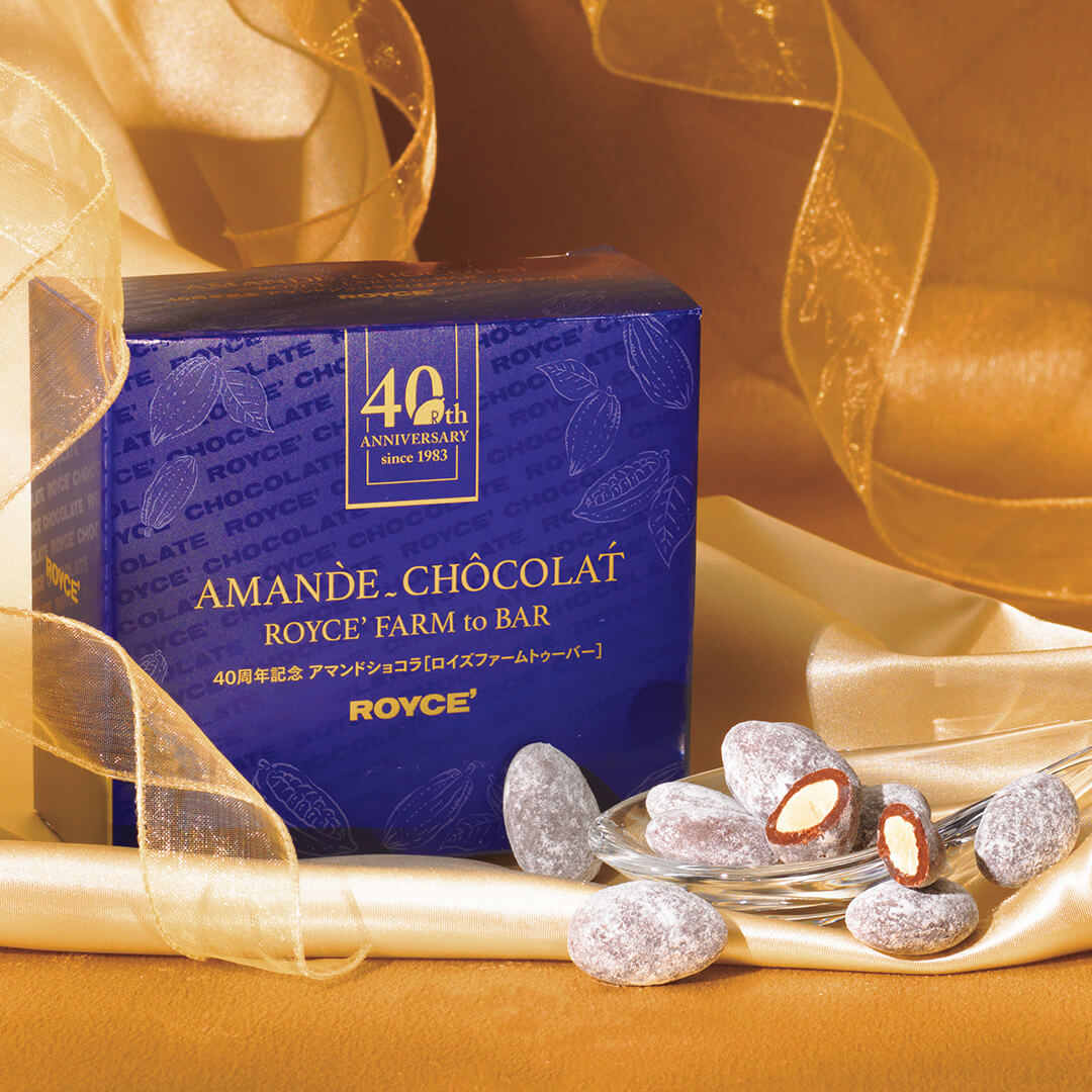 Image shows a crystal spoon with chocolate-coated almonds coated with white sugar powder. Blue box behind has text saying 40th Anniversary since 1983 Amande Chocolat ROYCE' Farm to Bar. ROYCE'. Background is in gold.