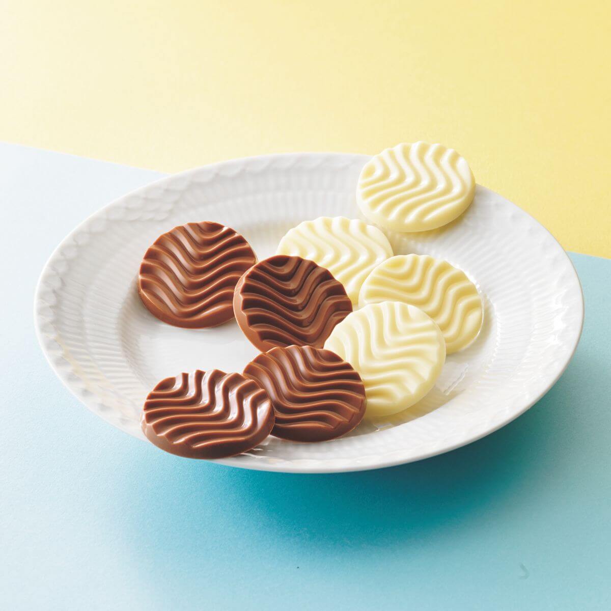 Pure Chocolate Creamy Milk & White features ripple-patterned chocolate discs on a white plate: four milk and four white, showcasing premium craftsmanship and flavor harmony. Background is in yellow and blue.