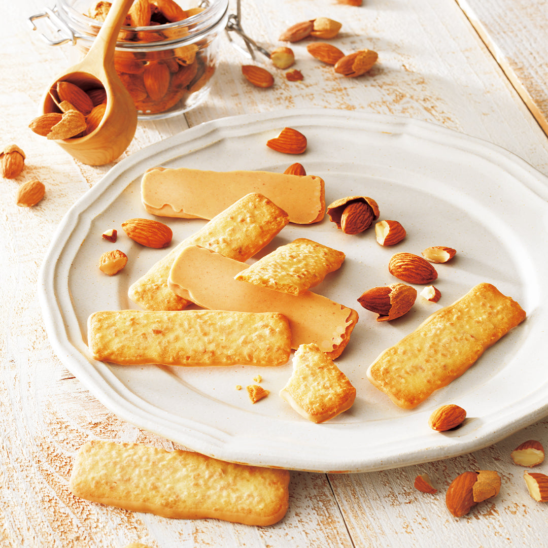 Baton Cookies "Almond" - Image shows almond cookies served on a white ceramic plate with scattered almonds and a small glass jar of almonds in the background, set on a light wooden table.
