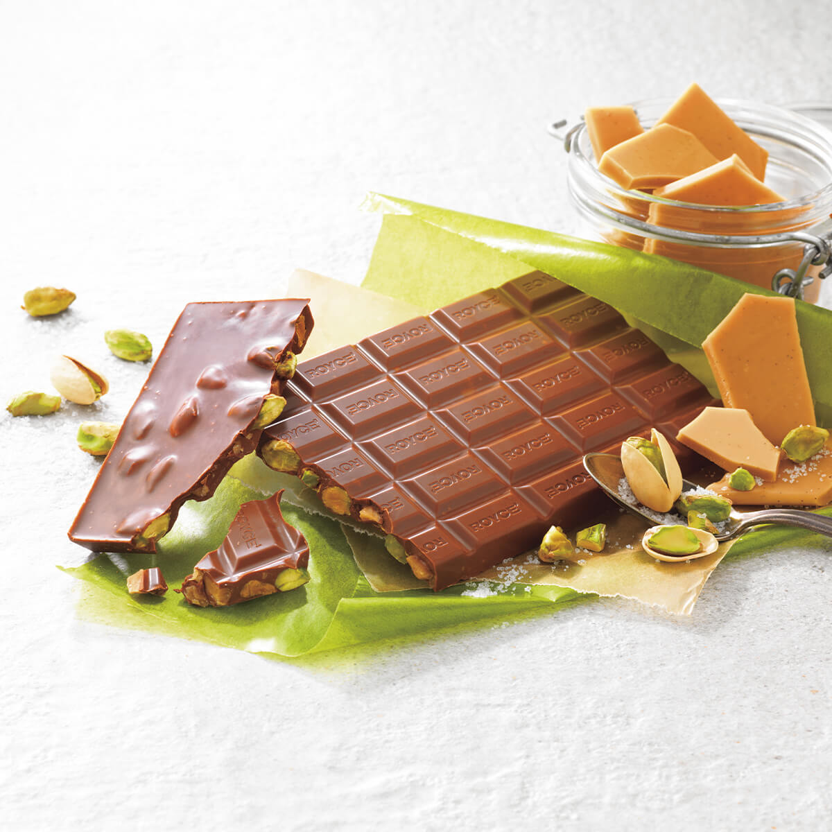 Chocolate Bar "Salted Pistachio & Toffee". Image shows a chocolate bar with almonds and toffee bits. Accents include a green paper, some nuts and toffees, and a spoon.