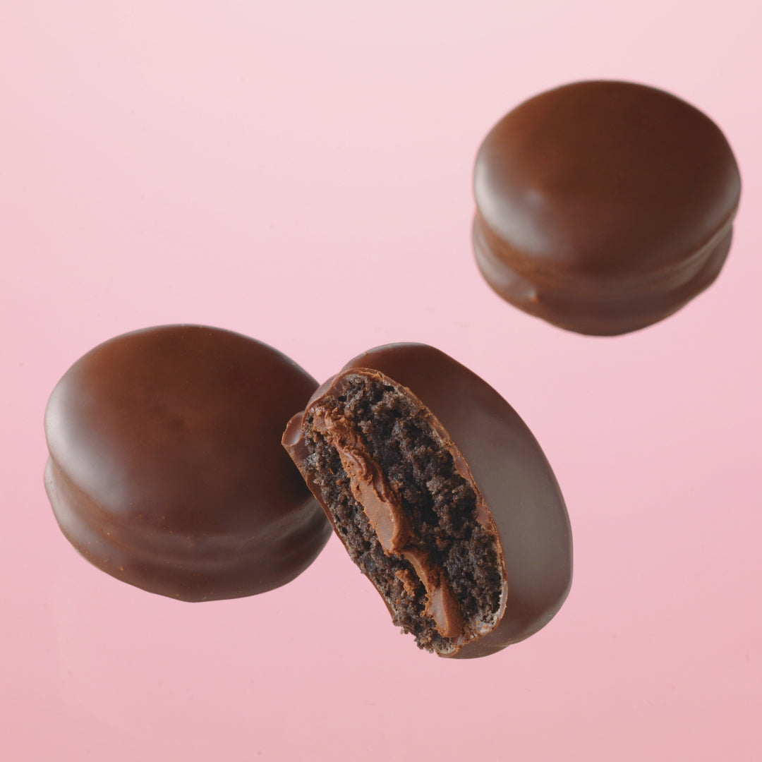 Cacao Macaron (6 Pcs) - Image shows chocolate-coated macarons. Background is in pink.