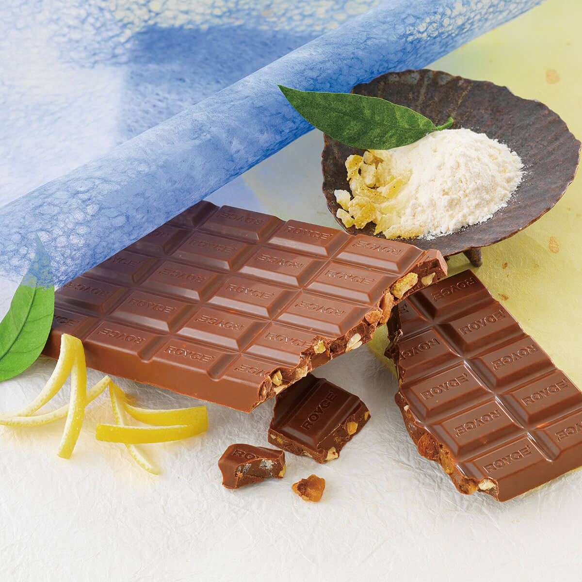 ROYCE' Kyoto Chocolate Bar "Wasanbon Toffee & Yuzu" - Image shows a chocolate bar with the words ROYCE' etched. Accents include a brown bowl with sugar and leaves, loose leaves and yuzu peels, and blue and yellow paper.