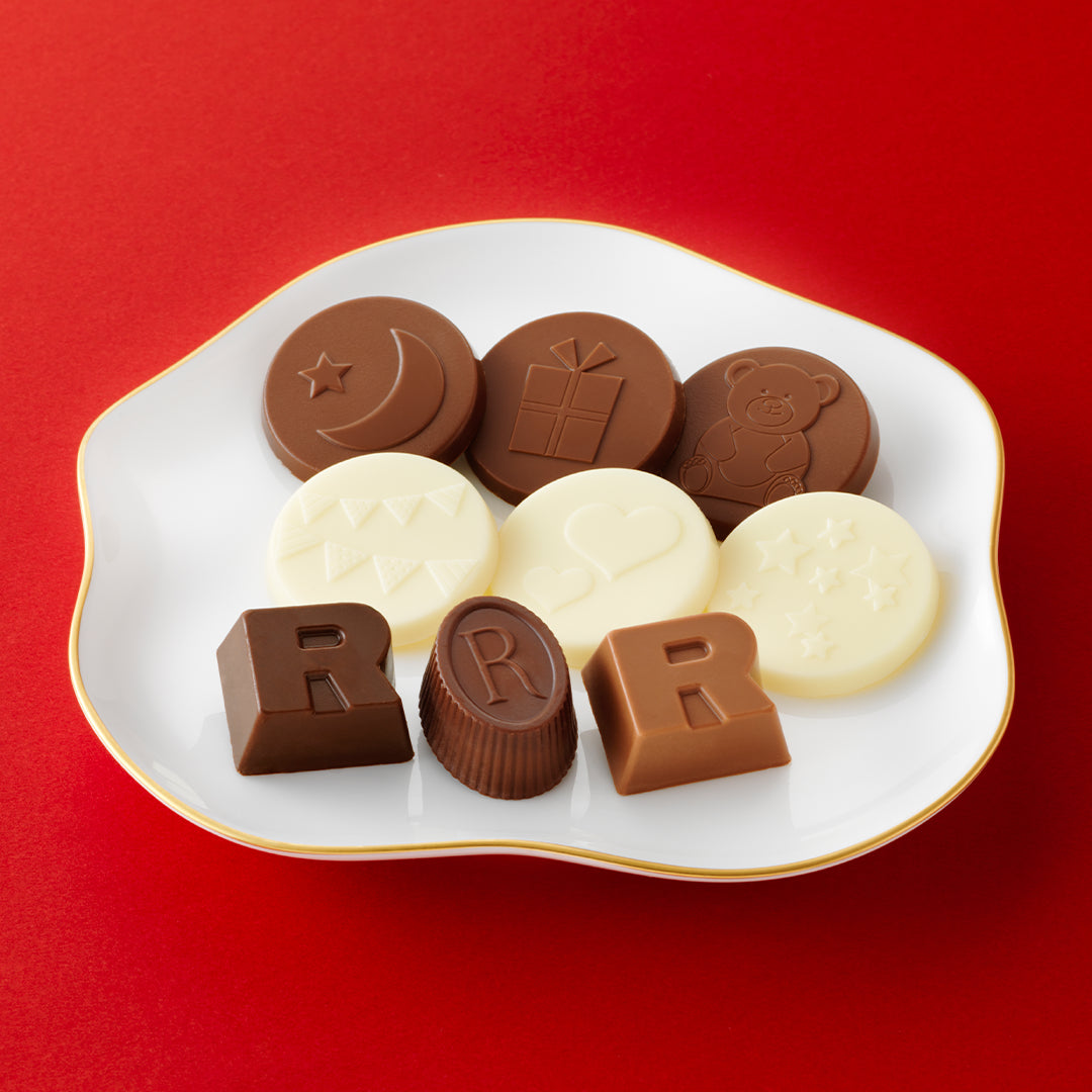 An assortment of chocolates arranged on a white scalloped plate with a gold rim, set against a solid red background. The chocolates include various shapes and colors: dark brown chocolates with embossed designs (moon and star, gift box, teddy bear), milk brown chocolates shaped like the letter R, and white milk chocolate pieces with embossed symbols.