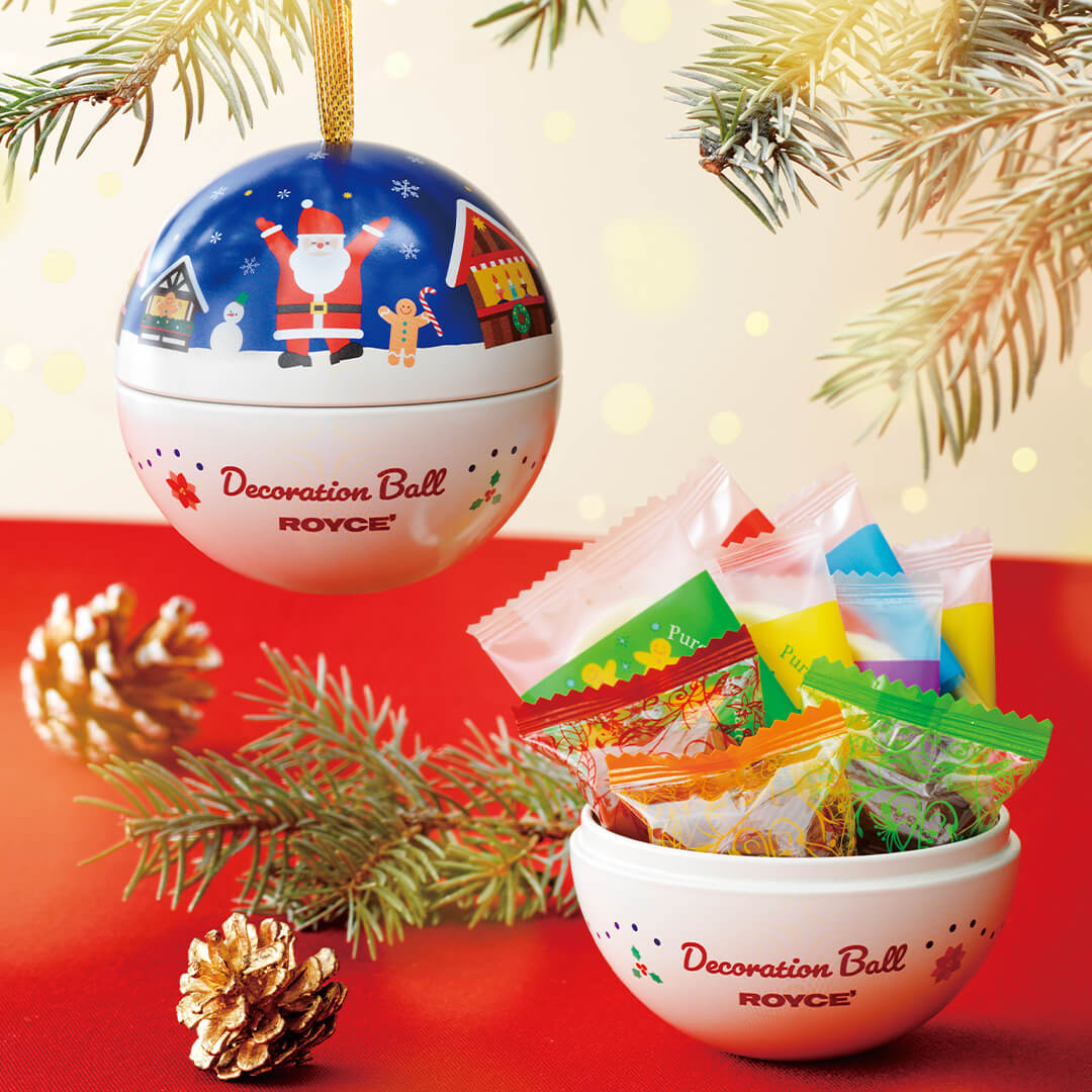 ROYCE' Chocolate - Decoration Ball "Christmas" - Image shows decorative orbs with blue and white colors and illustrations. Text says Decoration Ball ROYCE'. Accents include leaves, gilded acorns, and individually-wrapped chocolates. Background is in light gold and red.