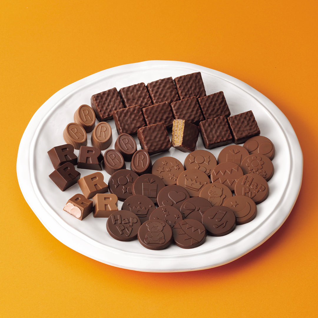ROYCE' Halloween Wakuwaku Box - An assortment of milk and dark chocolates arranged on a white oval plate against a solid orange background. The chocolates include waffle-textured squares, round cookies with embossed lettering, and a broken piece showing a biscuit layer.