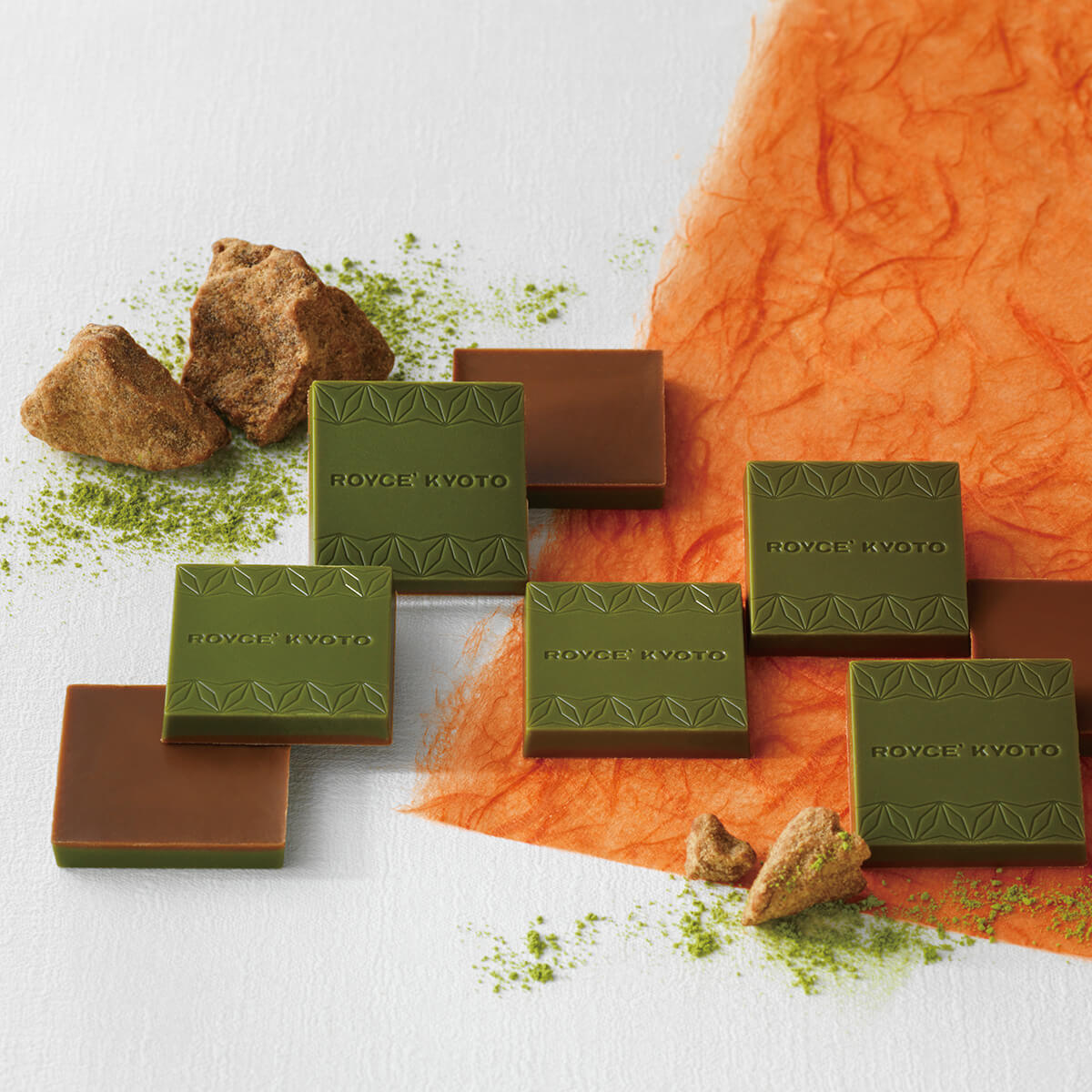 ROYCE' Kyoto Kokutoh & Uji Matcha Chocolate - Image shows brown and green chocolate blocks with the words ROYCE' Kyoto etched on them. Accents include loose matcha dustings and crumbles of kokutoh brown sugar. Surface is in white color with orange paper.