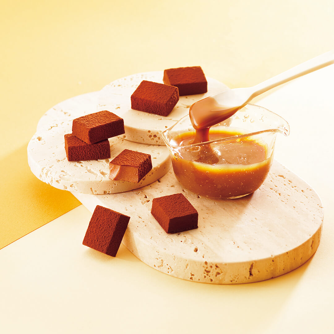 ROYCE' Chocolate - Nama Chocolate "Caramel Milk" - Image shows brown chocolate blocks on beige platforms. Accents include a spatula with caramel and a bowl filled with caramel.