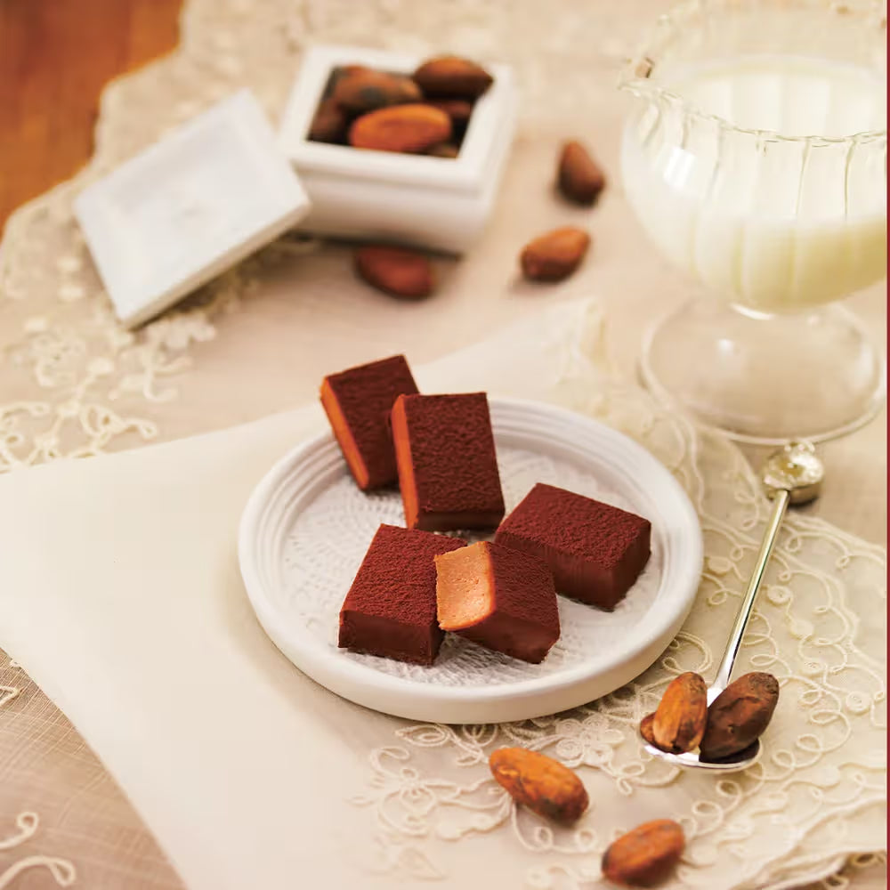 Nama Chocolate Mild Milk squares on a plate, cocoa-dusted, with one cut open, surrounded by cocoa beans and milk, reflecting its creamy, rich texture.