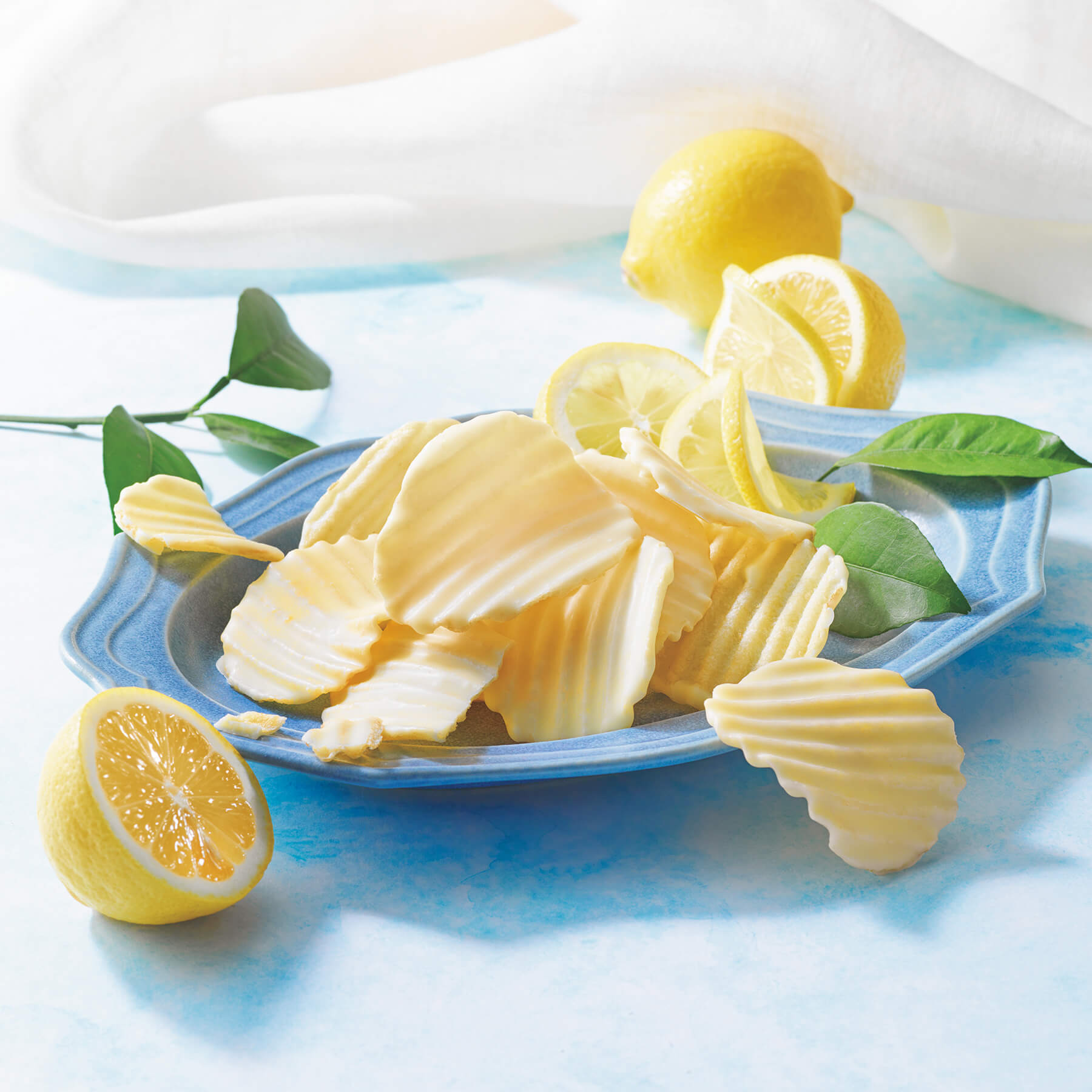ROYCE' Chocolate - Potatochip Chocolate "Lemon" - Image shows potato chips coated with white chocolate on a blue plate. Accents include yellow lemons and green leaves.