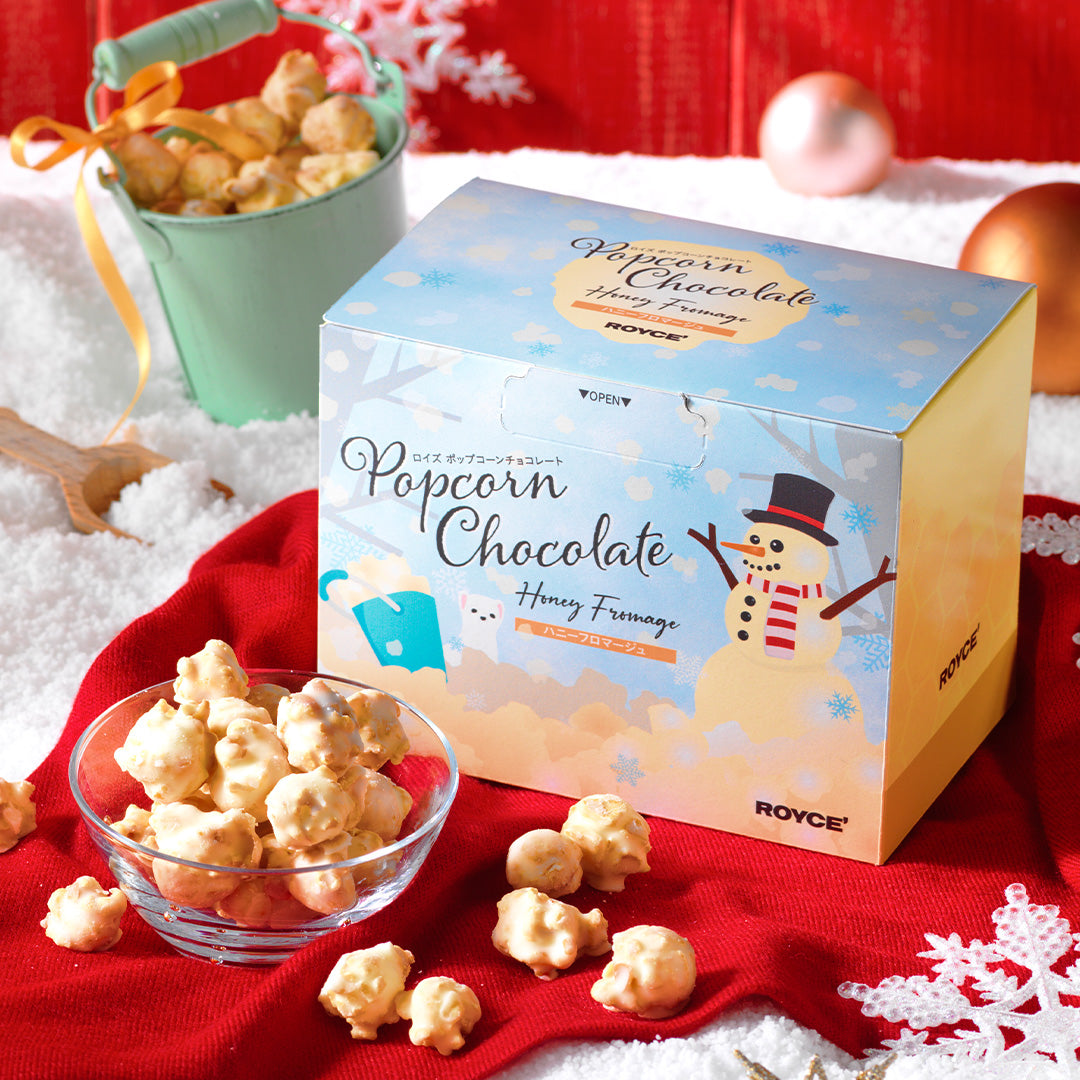 Image shows a printed box with snowman and other illustrations. Accents include loose popcorns, a clear bowl, a red cloth, a green pail, a gold ribbon, a wooden shovel, decorative balls, and paper cutouts.