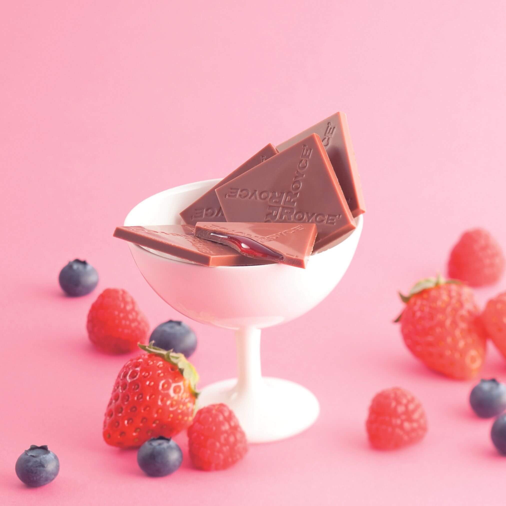 ROYCE' Chocolate - Prafeuille Chocolat "Berry Cube" - Image shows brown chocolate squares with red sauce and engraved with the words "ROYCE'", as placed in a white glass. Accents include red strawberries and raspberries and dark blue blueberries. Background is in pink color.