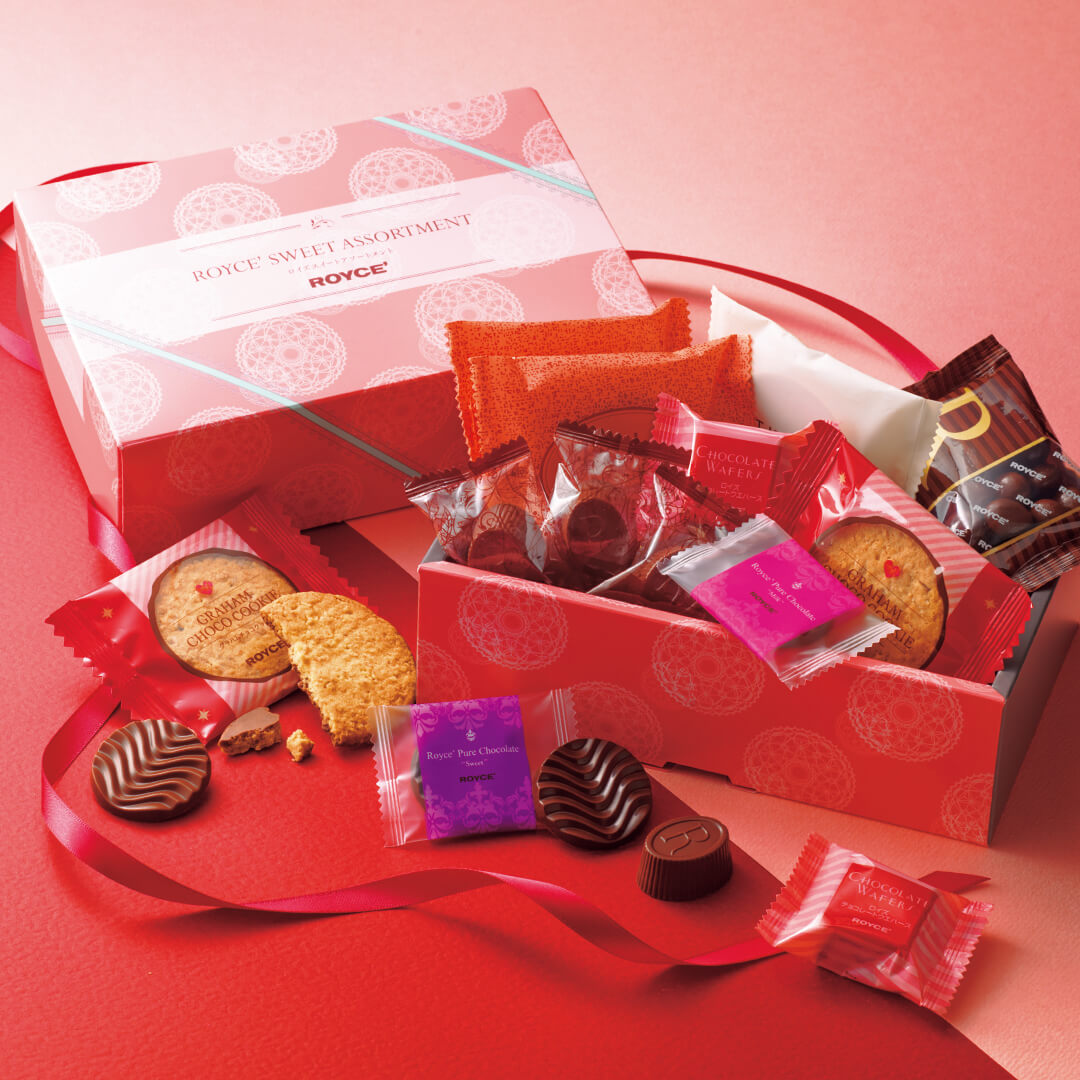 ROYCE' Chocolate - ROYCE' Sweet Assortment (17 Pcs) - Image shows two boxes on a red and pink surface with red ribbons. Box on top left is in the colors of white and pink with various prints and text saying ROYCE' Sweet Assortment ROYCE'. Box in bottom middle right shows a blue and pink printed box that is filled with individually-wrapped confections in various shapes, sizes, and colors.