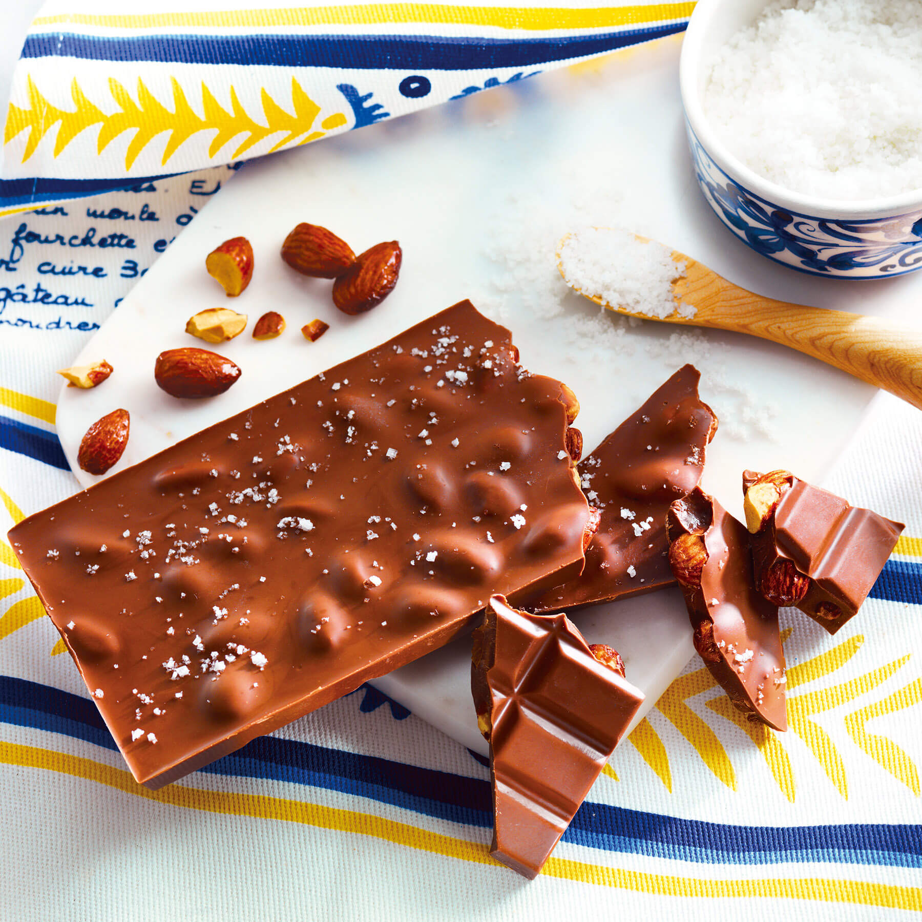 ROYCE' Chocolate - ROYCE' Tablette Chocolat "Amande Salée" - Image shows a brown chocolate bar with almonds and a sprinkling of salt, as placed on a white plate with accents of a wooden spoon, loose salt, and a cup filled with salt. There is also a placemat with various prints in yellow, blue, and white.