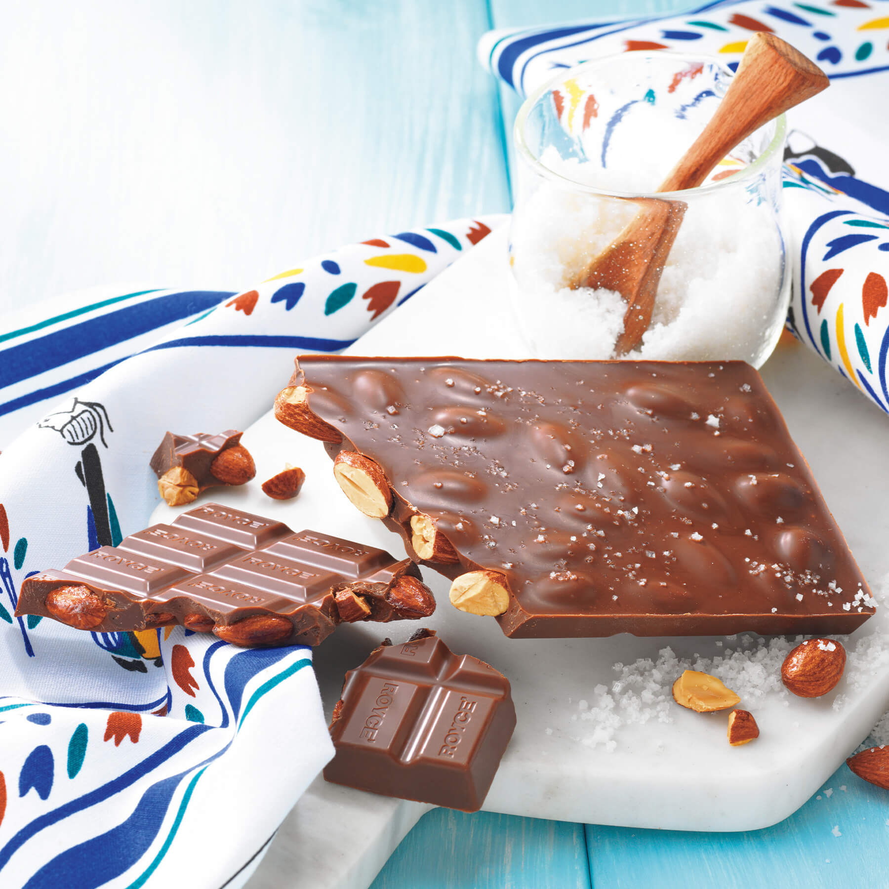 ROYCE' Chocolate - ROYCE' Tablette Chocolat "Amande Salée" - Image shows a brown chocolate bar with almonds and a sprinkling of salt, as placed on a white plate with accents of loose salt, a glass filled with salt and a wooden spoon, and a placemat in various prints in the colors of white, black, green, brown, blue, and yellow. Background is in wooden blue.
