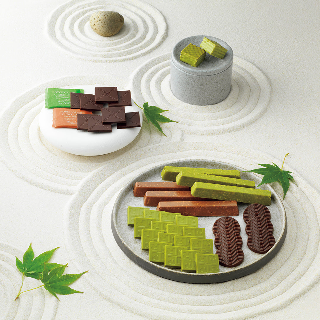 ROYCE' Variety Pack "Flavorful" - An assortment of Japanese sweets, including green and brown rectangular bars and chocolate pieces, arranged on ceramic plates over a white surface with circular raked garden-style sand patterns and green leaves.