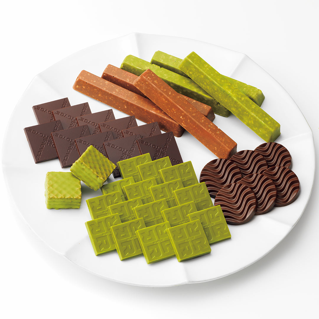 ROYCE' Variety Pack "Flavorful" - An assortment of chocolates and candy bars arranged on a white plate against a clean white background. The assortment includes dark brown chocolate bars, orange-brown stick bars, light green stick bars, green square chocolates, and wavy dark brown chocolate pieces.