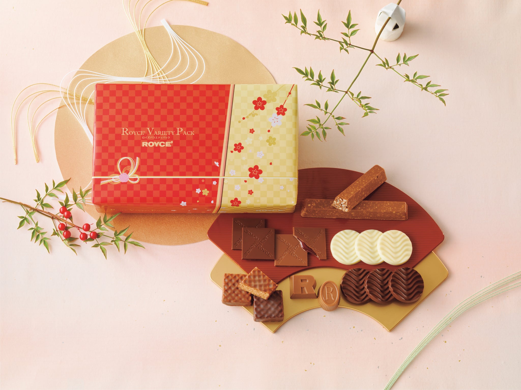 Image shows a red and gold box with checkers and floral prints. Text says ROYCE' Variety Pack ROYCE'. Accents include gold and brown trays with chocolates of varying sizes and shapes in the colors brown and white. There are also white, green, and gold threads together with white paper origami and brown and green twigs with green leaves and red berries. Background is in light pink.