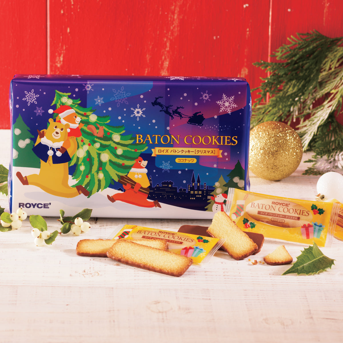 Image shows a blue box with illustrations of snowflakes, bears, trees, a cityscape, and a snowman. Text says Baton Cookies ROYCE'. Accents include baton-shaped cookies, some with wrapping saying ROYCE' Baton Cookies. Other accessories include green leaves, white bulbs, and a golden ball. Background is in red and surface is in white.