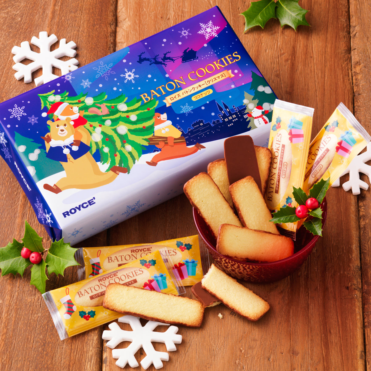 Image shows a blue box with illustrations of snowflakes, bears, trees, a cityscape, and a snowman. Text says Baton Cookies ROYCE'. Accents include mistletoes in green and red and white snowflakes. Other accessories include a red bowl filled with baton-shaped cookies, some with wrapping saying Baton Cookies. Surface is brown wood.