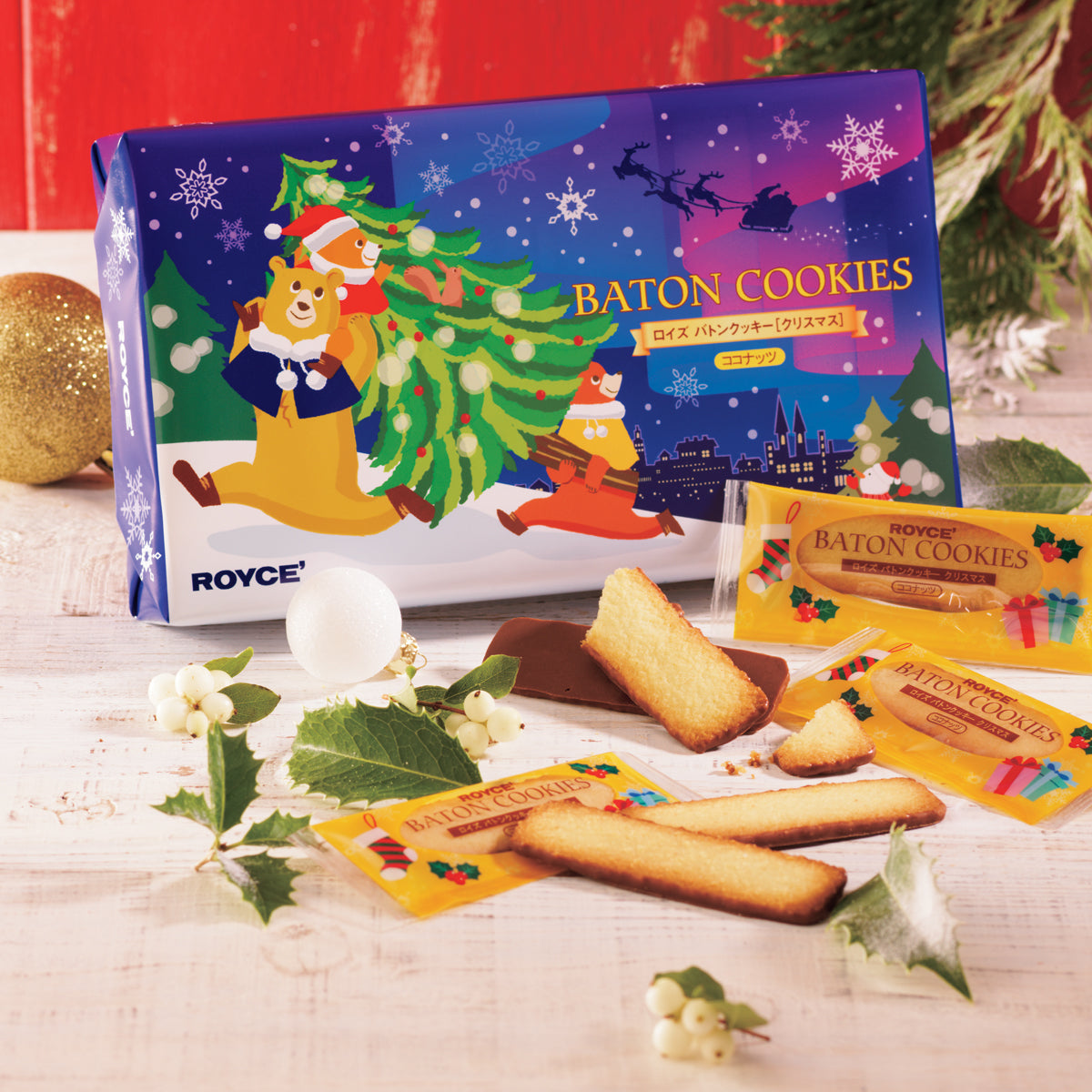 Image shows a blue box with illustrations of snowflakes, bears, trees, a cityscape, and a snowman. Text says Baton Cookies ROYCE'. Accents include baton-shaped cookies, some with wrapping saying ROYCE' Baton Cookies. Other accessories include green leaves, white bulbs, and a golden ball. Background is in red and surface is in white.