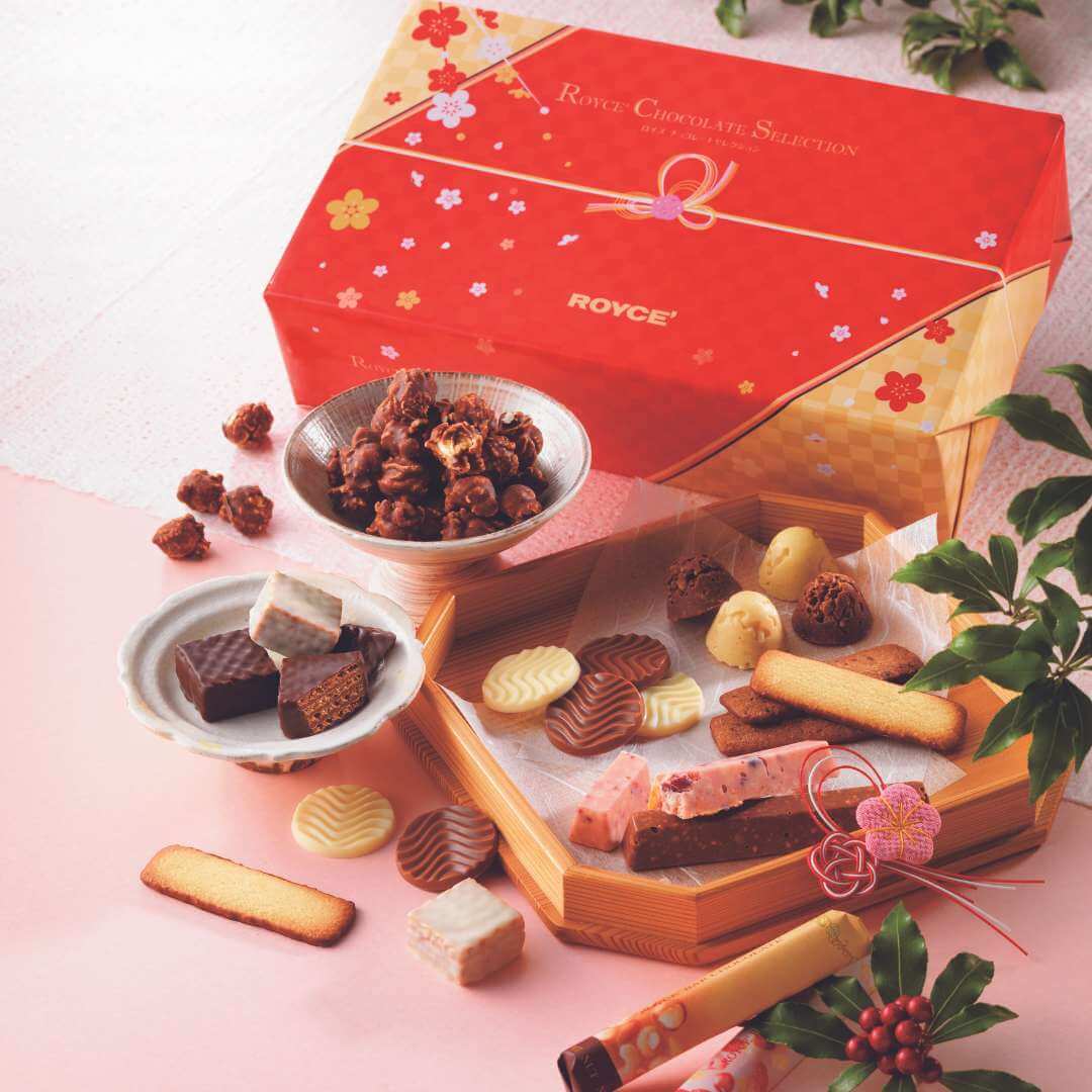 Image shows a red box with gold edges and floral prints. Text on box says ROYCE' Chocolate Selection ROYCE'. Accents include wooden trays and ceramic bowls with different chocolates and confections in varying shapes, sizes, and colors. There are also some green leaves on the right. Background is pink.