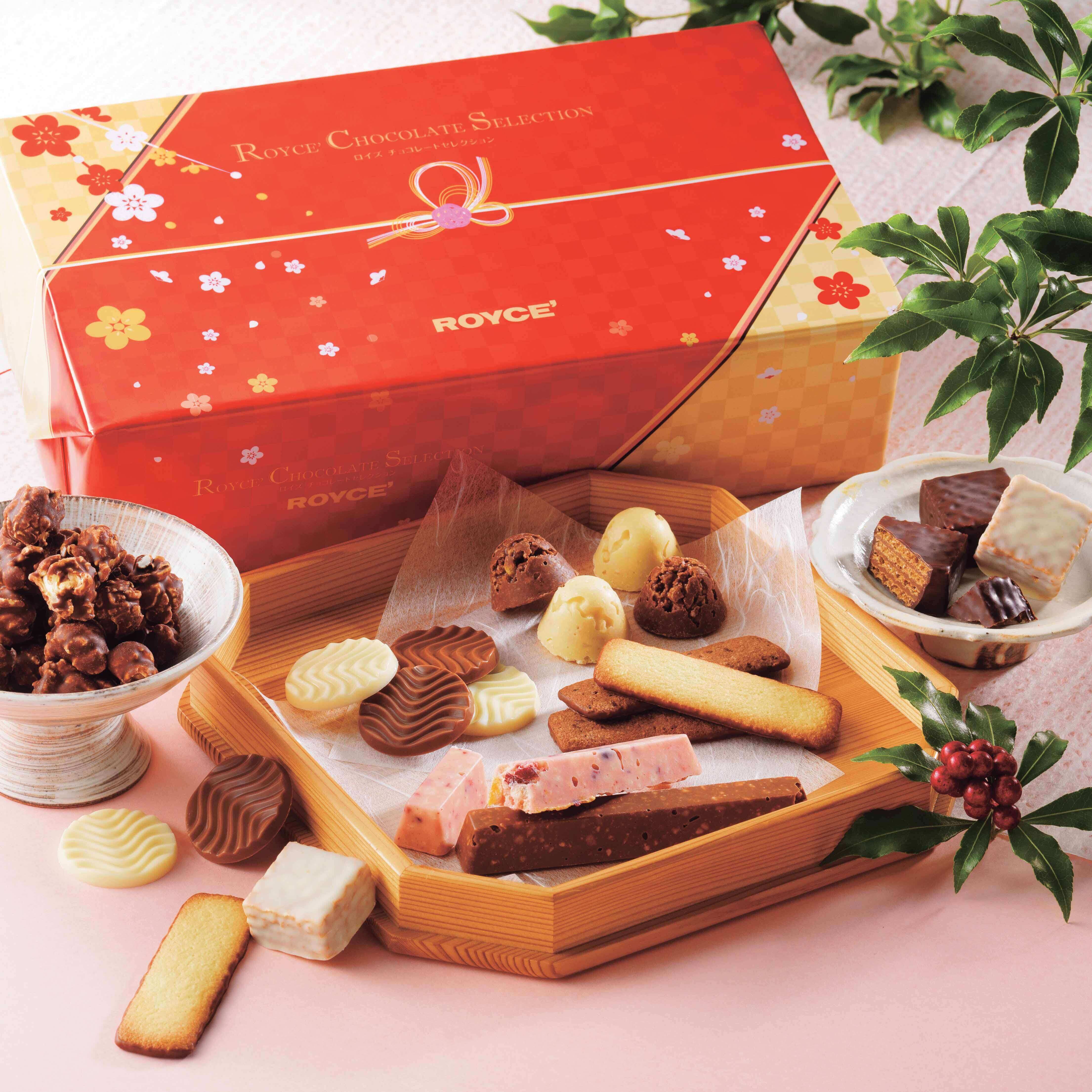 Image shows a red box with golden edges and floral prints on a pink table. Accents include wooden trays and ceramic bowls with different chocolates and confections in varying shapes, sizes, and colors. There are also some green leaves and red berries on the right.