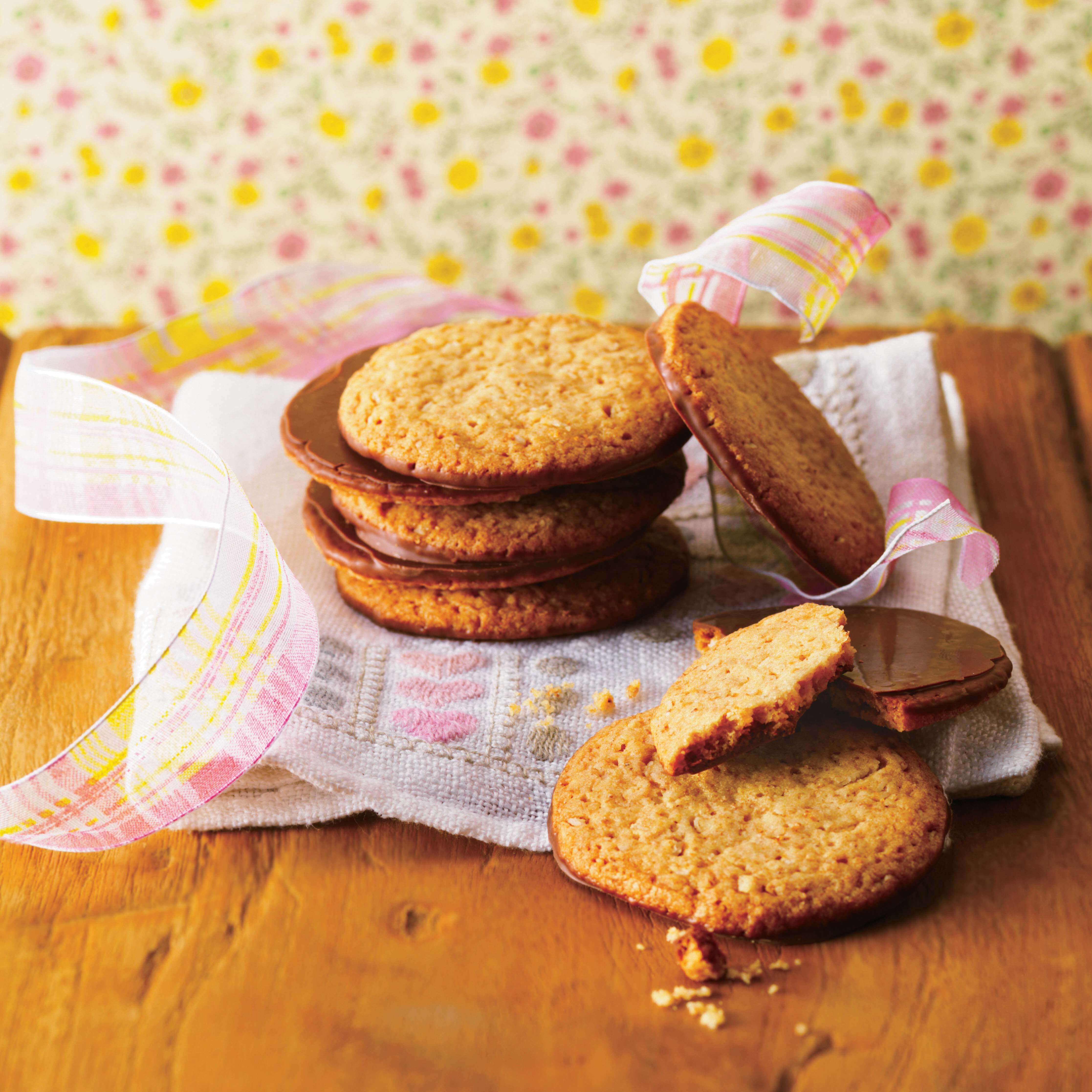 ROYCE' Chocolate - Graham Chocolate Cookie - Image shows chocolate-coated cookies on a wooden surface with a plaid ribbon and embroidered towel. Background has blurry floral prints.