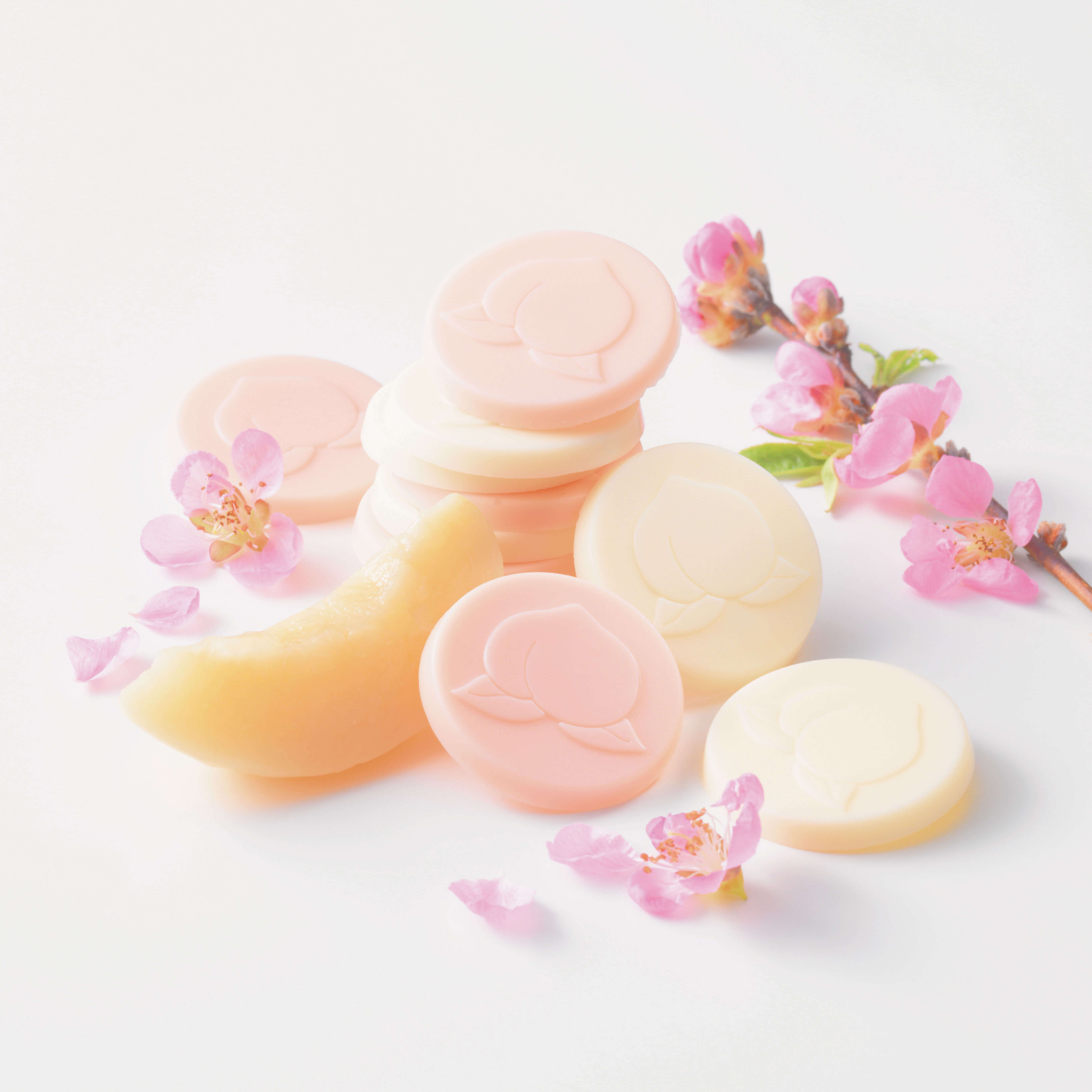 ROYCE' Chocolate - Hakutouberry & Hakutouwhite Chocolate - Image shows light pink and white chocolate discs embossed with a peach-shaped pattern, as placed on a white surface. Accents include a slice of peach, pink flowers, and a brown twig with green leaves and pink flowers.