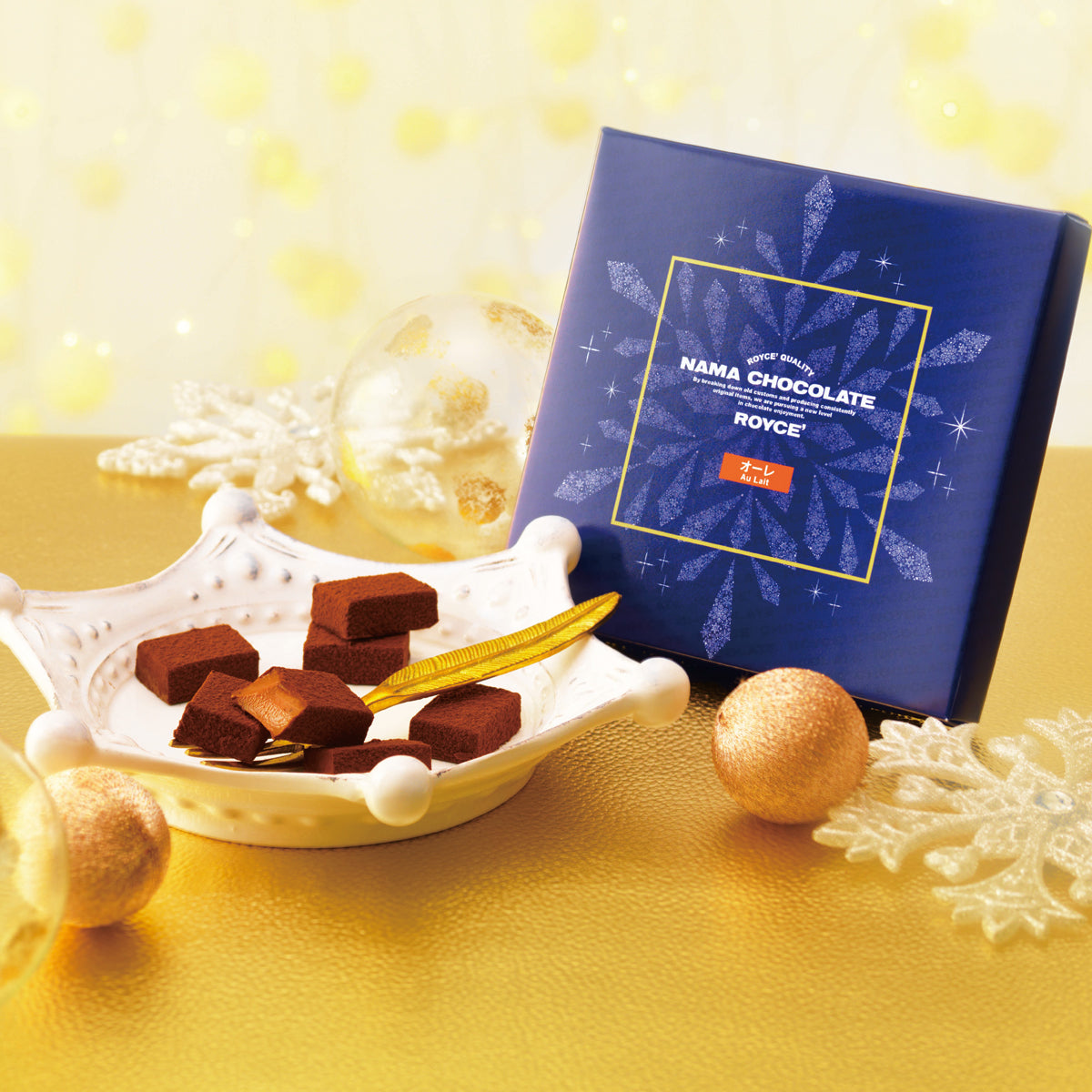 Image shows brown chocolate blocks on a white plate with golden leaf. Blue box with snowflake and gold square prints on right has text saying ROYCE' Quality Nama Chocolate ROYCE' Au Lait. Accents include golden balls and snowflakes. Background and surface are both in gold color.