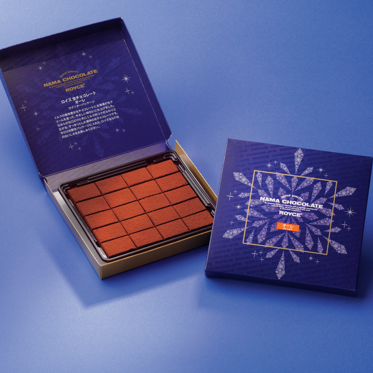 Image shows a blue box on the right printed with the words ROYCE' Chocolate and has illustrations of a snowflake and stars in white and golden square in the middle. Text says ROYCE' Quality Nama Chocolate ROYCE' Au Lait. Blue box on left has brown chocolate blocks. Text says ROYCE' Quality Nama Chocolate ROYCE'. Background surface is in blue.