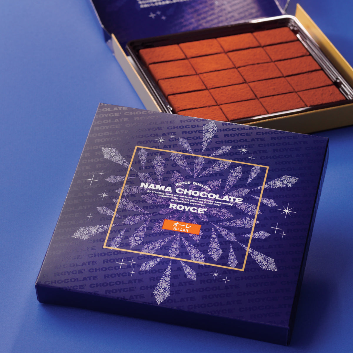 Image shows a blue box printed with the words ROYCE' Chocolate and has illustrations of a snowflake and stars in white and golden square in the middle. Text says ROYCE' Quality Nama Chocolate By breaking down old customs and producing consistently original items, we are pursuing a new level in chocolate enjoyment. Au Lait. In the background is a blue and gold box with brown chocolate blocks. Background surface is in blue.