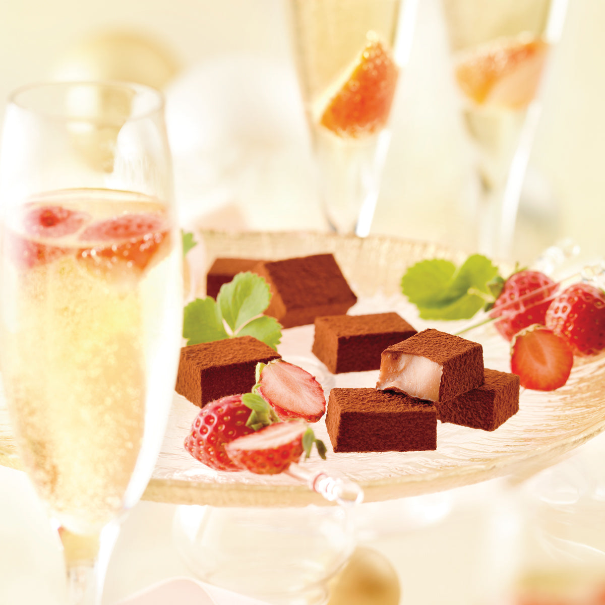 Nama Chocolate Strawberry Champagne squares on a glass platter, dusted with cocoa, surrounded by fresh strawberries, with champagne flutes in the background.