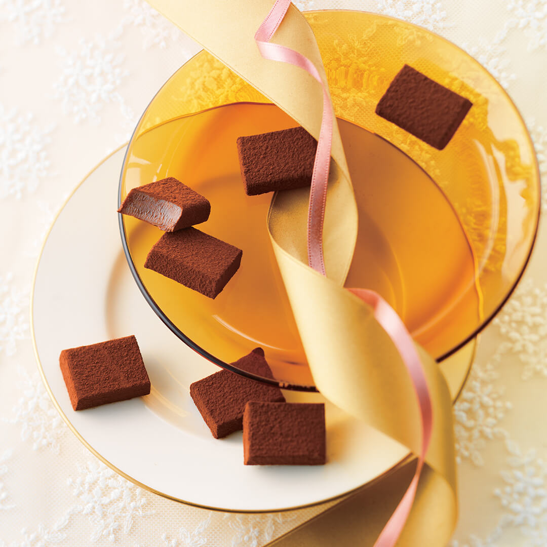 ROYCE' Chocolate - Nama Chocolate "Caraméliser" - Image shows brown chocolate blocks on white and gold plates. Accents include gold and pink ribbons, and a tablecloth with lace detail.