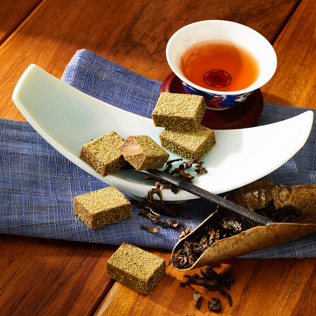 ROYCE' Chocolate - Nama Chocolate "Oolong Tea" - Image shows chocolate blocks that have a light brown color with green powder on top. The chocolate blocks are placed on a crescent-shaped white plate. Accents include a blue tablecloth, a red plate and printed tea cup with amber-hued tea inside, a silver fork with black handle, and a leaf-shaped bronze plate with loose black tea leaves and shavings. Background is a wooden surface in brown.