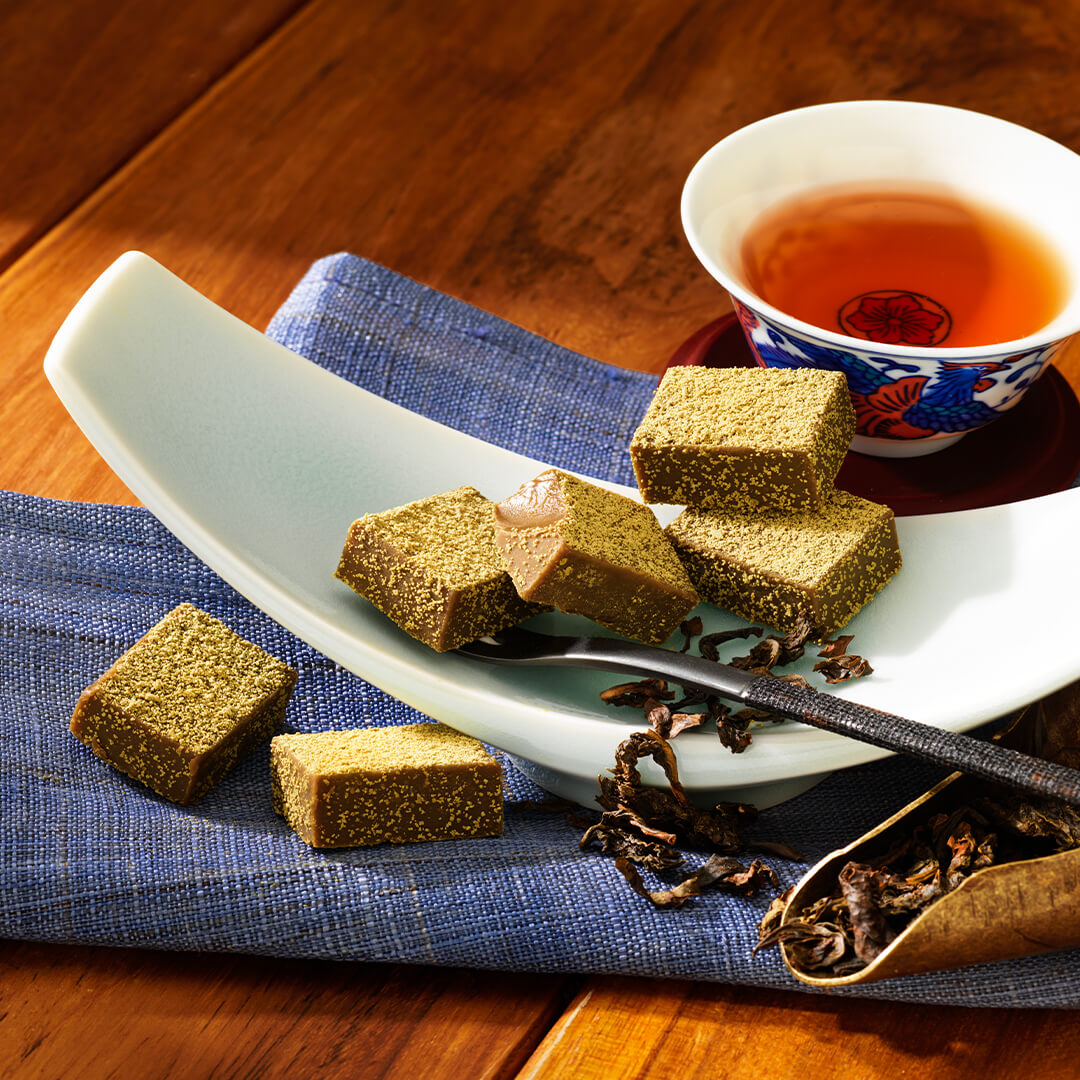 ROYCE' Chocolate - Nama Chocolate "Oolong Tea" - Image shows chocolate blocks that have a light brown color with green powder on top. The chocolate blocks are placed on a crescent-shaped white plate. Accents include a blue tablecloth, a red plate and printed tea cup with amber-hued tea inside, a silver fork with black handle, and a leaf-shaped bronze plate with loose black tea leaves and shavings. Background is a wooden surface in brown.