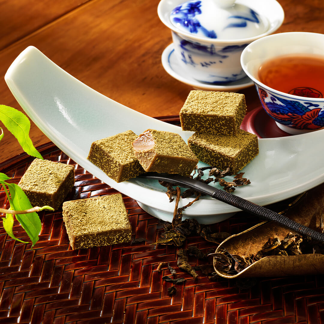 ROYCE' Chocolate - Nama Chocolate "Oolong Tea" - Image shows chocolate blocks that have a light brown color with green powder on top. The chocolate blocks are placed on a crescent-shaped white plate and a brown wooden plate with basket weave pattern. Accents include a blue tablecloth, a white tea set with prints, green leaves, a red plate and printed tea cup with amber-hued tea inside, a silver fork with black handle, and a leaf-shaped bronze plate with loose black tea leaves and shavings.