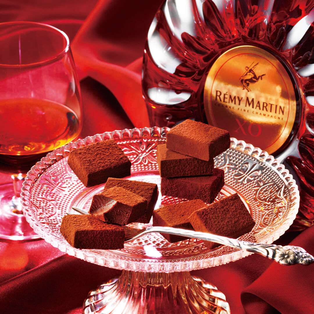 Image shows brown chocolate blocks on a clear plate with stand and silver fork. Accents include a clear glass with amber-hued liquid and a circular bottle with amber-hued liquid. There is a golden disc on the bottle with a text that says Rémy Martin Cognac Fine Champagne XO. Background is in red with a crumpled cloth effect.