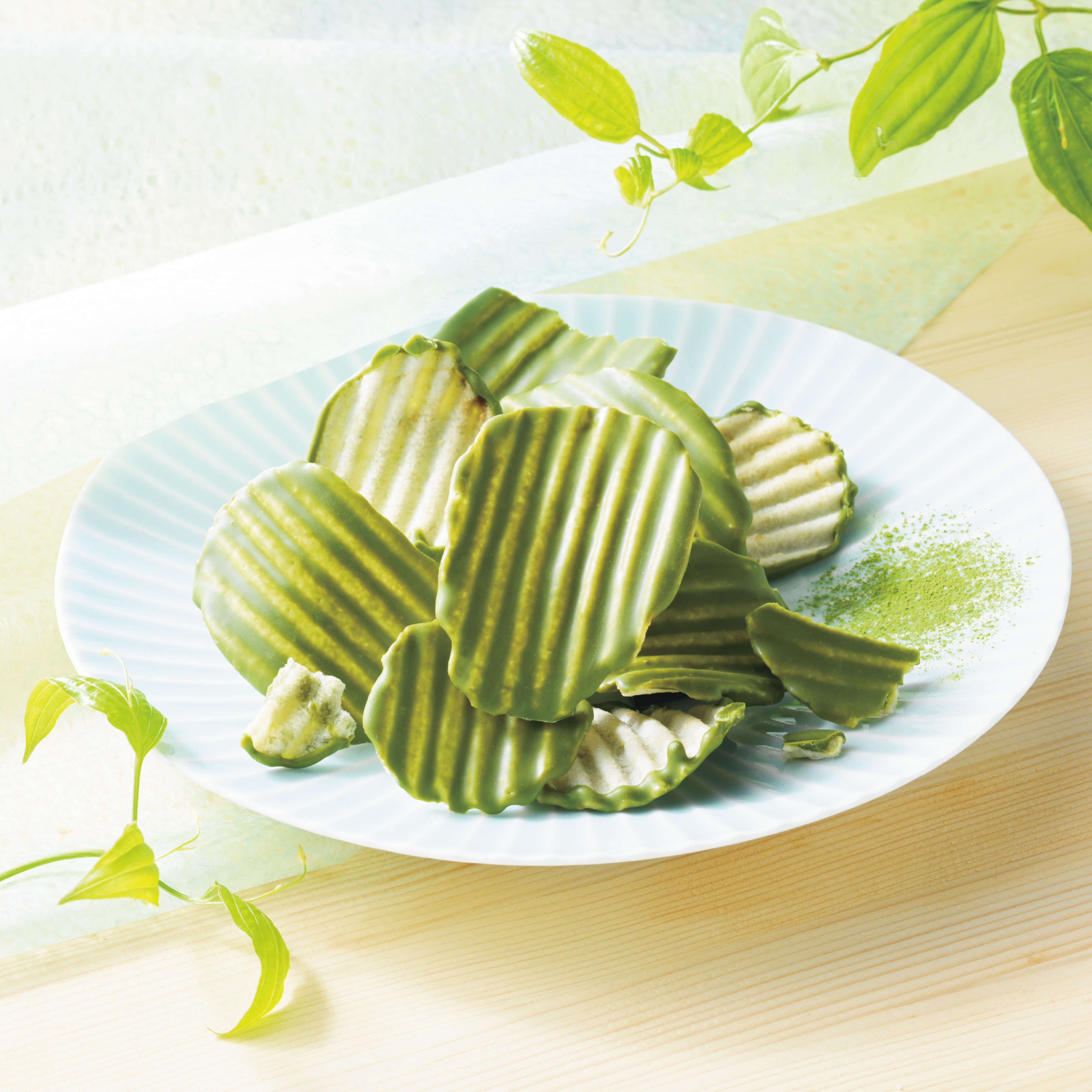 ROYCE' Chocolate - Potatochip Chocolate "Matcha" - Image shows green-colored potato chips on a plate with some matcha powder, as placed on a wooden surface. Accents include a green paper and some leaves.