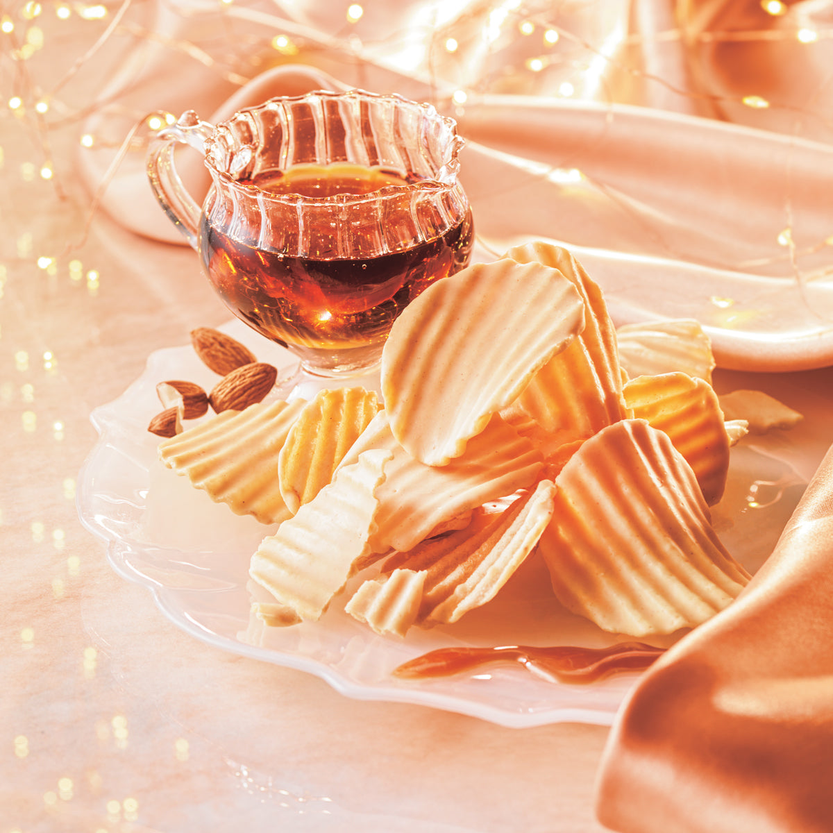 Image shows golden, ridged chocolate-coated potato chips on a clear plate with almonds and a cup with maple syrup. Accents include golden lights and a peach-colored tablecloth. Surface is in light peach.