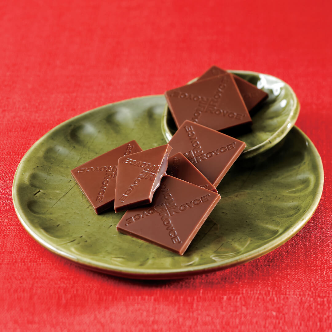 Image shows brown chocolate squares with the words ROYCE' engraved on them. Accents include two green round plates. Background is in red.