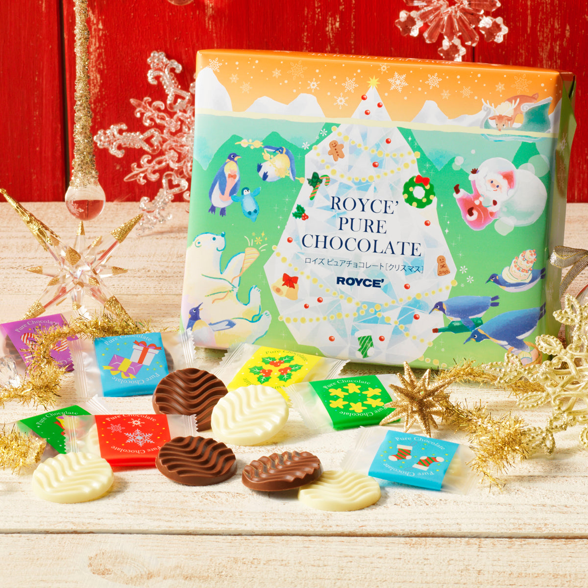 Image shows a green and orange box with illustrations of icebergs, animals, Santa Claus, plants, snowflakes, and holiday ornaments. Text says ROYCE' Pure Chocolate ROYCE'. Accents include golden and clear stars, snowflakes, ribbons, fuzzy trail, and a drop-like glass with gold glitters. Other accents include chocolate discs in brown and white with individually-wrapped chocolates in different colors. Background is in red and surface is in white.