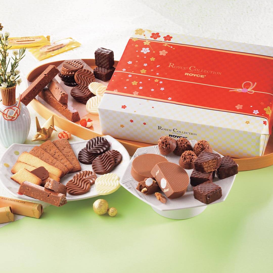 ROYCE' Chocolate - ROYCE' Collection "New Year" - Image shows a printed box in white and red with floral prints. Text on the box says ROYCE' Collection ROYCE'. Accents include wooden and ceramic plates filled with confections of varying colors and shapes. Other accessories seen are a white vase with flowers and some gold orbs and paper figurines. Background is a mix of white and green.