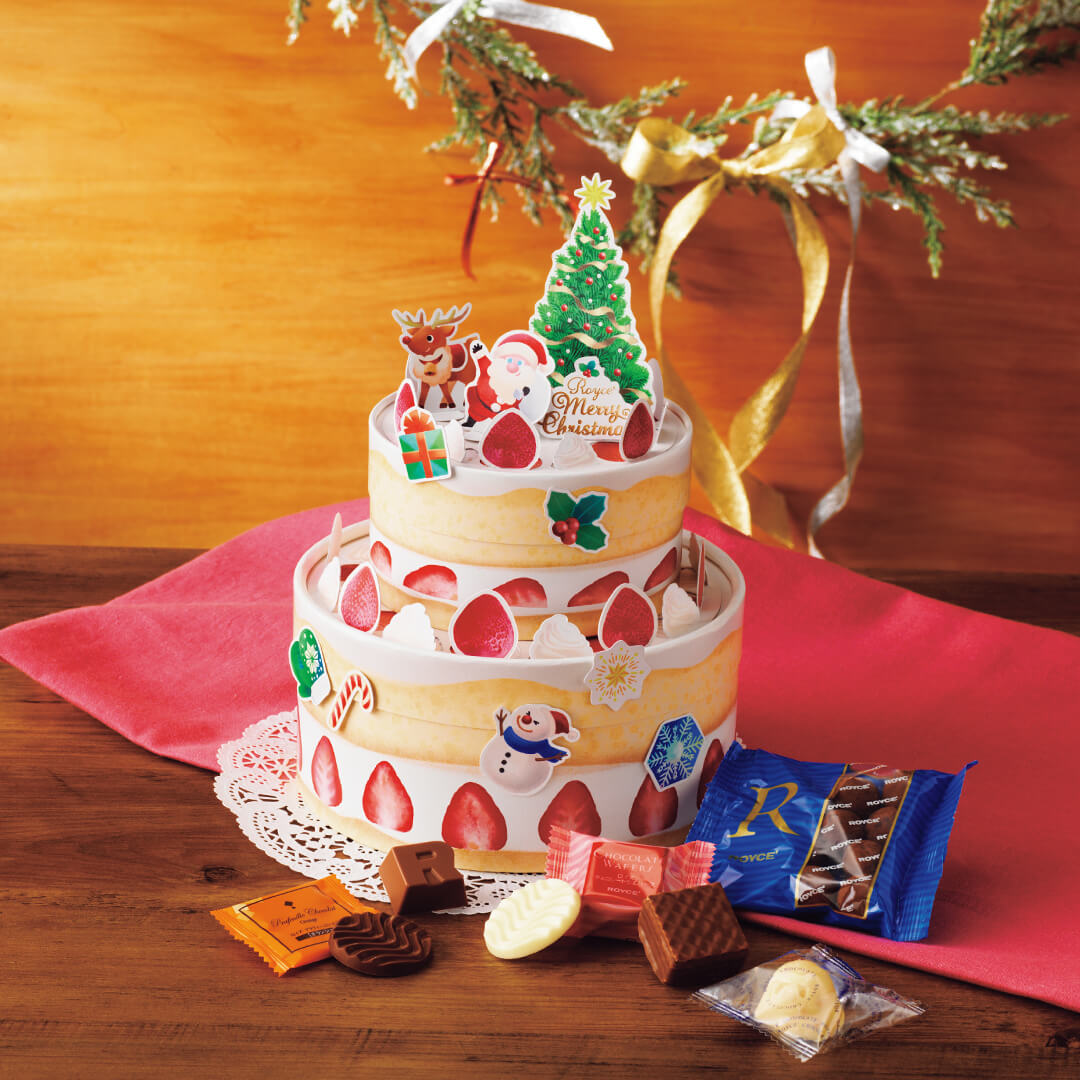 Image shows boxes that look like a two-tiered cake in yellow and white with accents like red strawberries, white whipped cream, a green Christmas tree, a Santa Claus, a reindeer, and other holiday-themed accents. Also seen in the picture are a red tablecloth, a white laced placemat, a green wreath with silver, gold, and red ribbons. A mix of loose confections in different shapes and colors can be seen too. Background is in dark yellow and light brown.