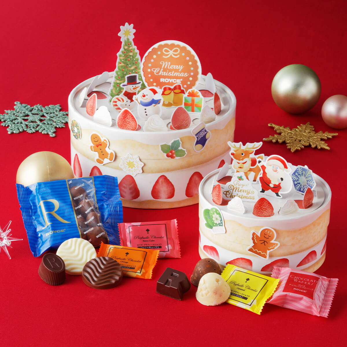 Image shows boxes that look like cakes in yellow and white with accents like red strawberries, white whipped cream, a green Christmas tree, a Santa Claus, a reindeer, and other holiday-themed accents. Also seen in the picture are bronze and rose gold orbs, a clear plastic star, and a snowflake-shaped cutouts in green and gold. A mix of loose confections in different shapes and colors can be seen too. Background is in red.