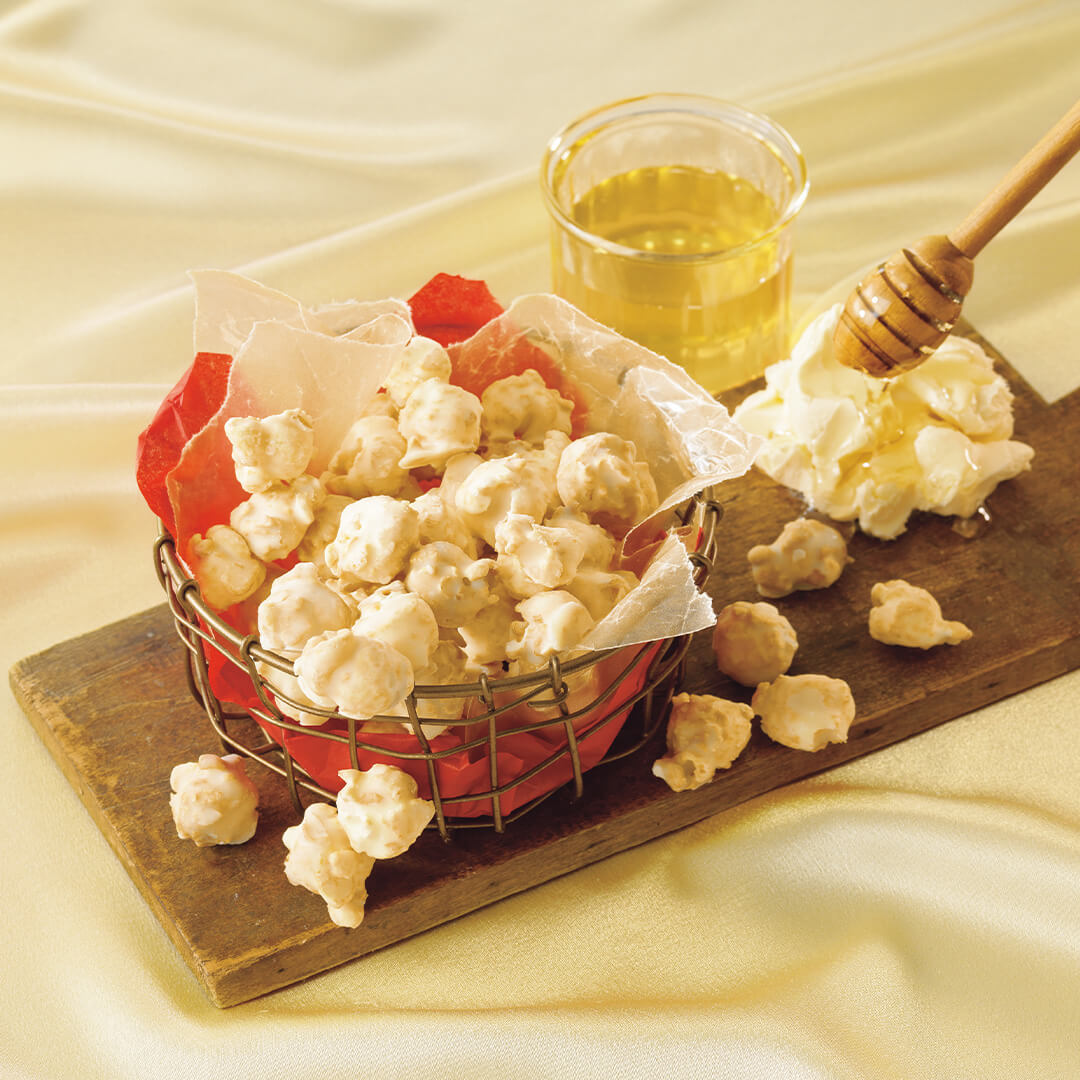 Image shows popcorn coated with white chocolate on a brown chopping board and a netted gold bowl with crumpled red and white paper. Accents include a scoopful of butter in yellow, a bowl filled with yellow honey, and a honey spool in brown. Background is in color light gold with a crumpled silk effect.