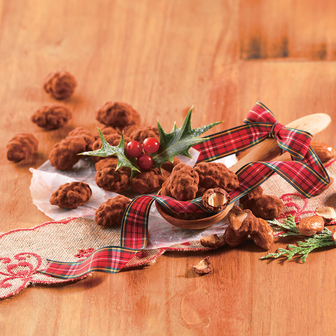 Image shows clusters of chocolate-covered almonds on a brown wooden table and a brown wooden spoon. Accents include a printed red ribbon with stripes in green, yellow, white, and black. Accents include green leaves and red berries, together with a white crinkled paper and a flesh-colored tablecloth with red trimmings. Background is in light brown.