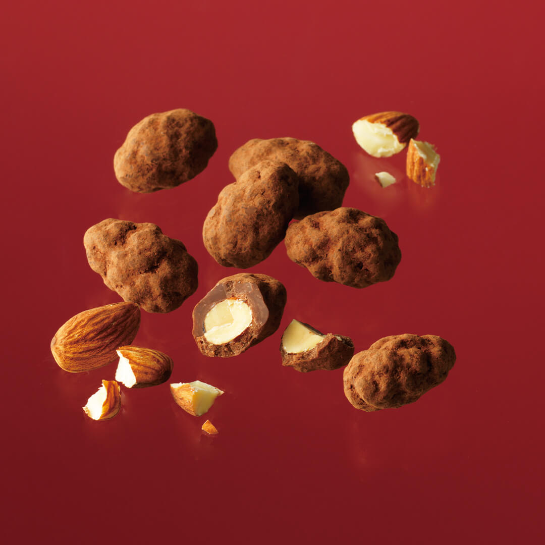 Image shows brown clusters of chocolate-coated almonds with an uneven surface and some loose almonds. Background is in red.