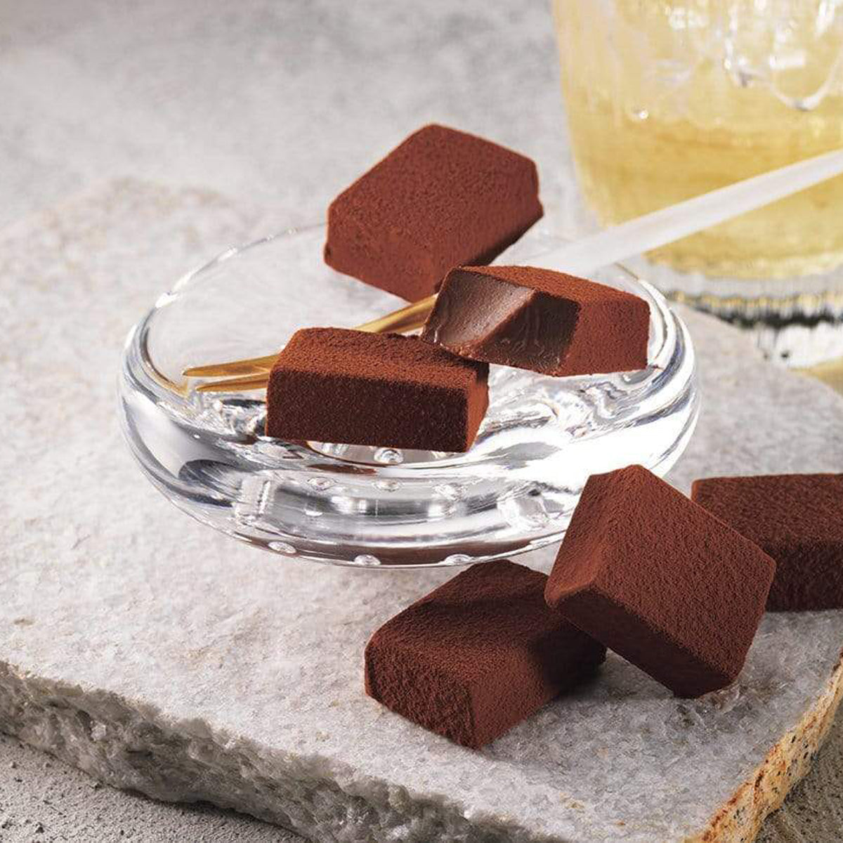 ROYCE' Chocolate - Nama Chocolate "Islay Whisky (Port Charlotte)" - Image shows brown blocks of chocolates on a clear plate and gray stone platform. Accents include a wooden stick and gray background shows a blurred picture of a glass with light yellow whisky.